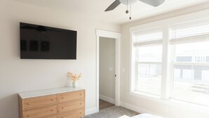 Un dormitorio con cama, cómoda, televisor montado en la pared y ventana con persianas.
