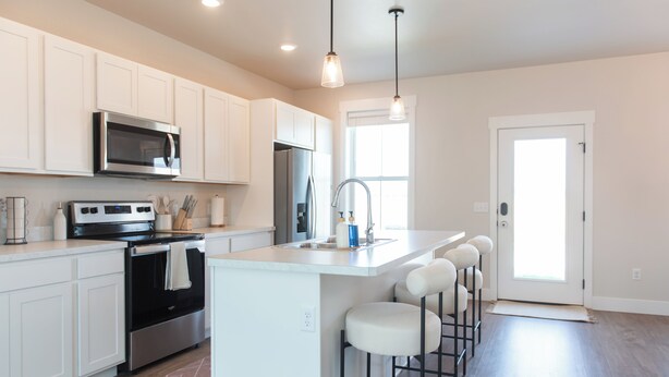 Una cocina moderna con armarios blancos, electrodomésticos de acero inoxidable, una isla central con taburetes y luminarias colgantes.
