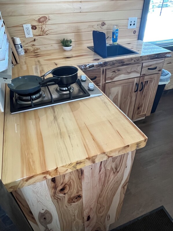 Une cuisine avec un plan de travail en bois, une cuisinière à gaz, une poêle, une petite plante en pot et une chaise bleue.