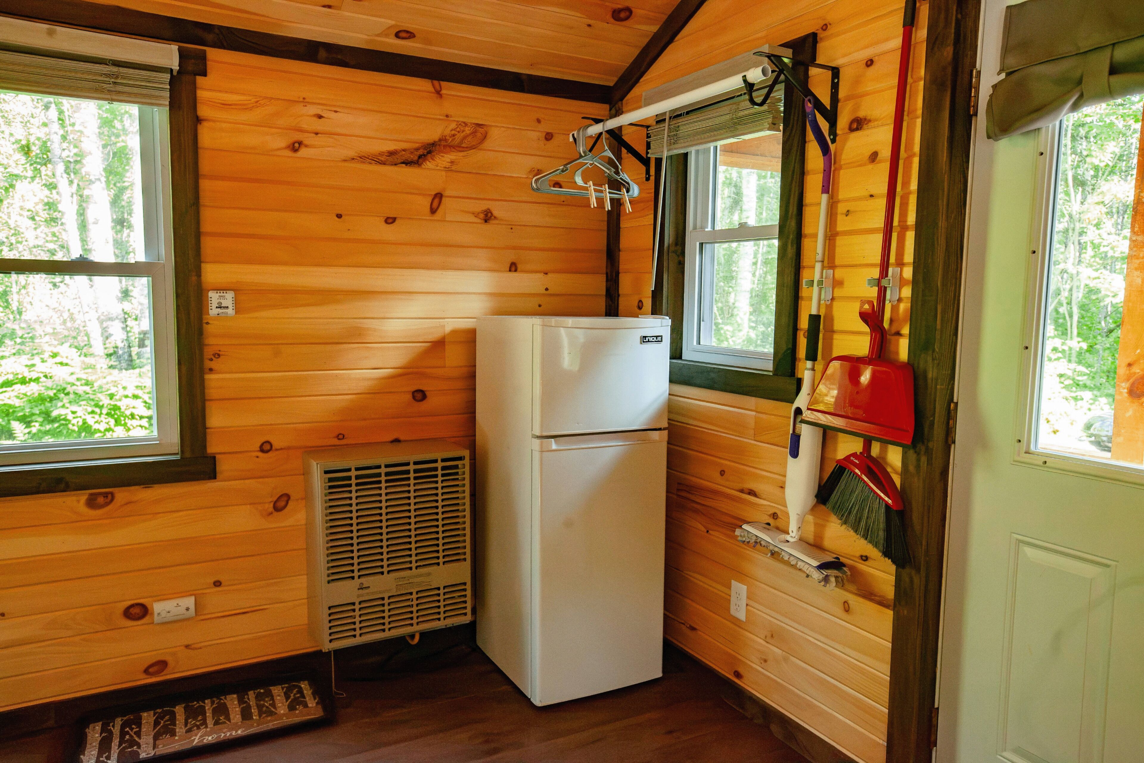 Une cuisine compacte aux murs en bois, avec un réfrigérateur blanc, une fenêtre donnant sur un paysage verdoyant et un balai accroché au mur.