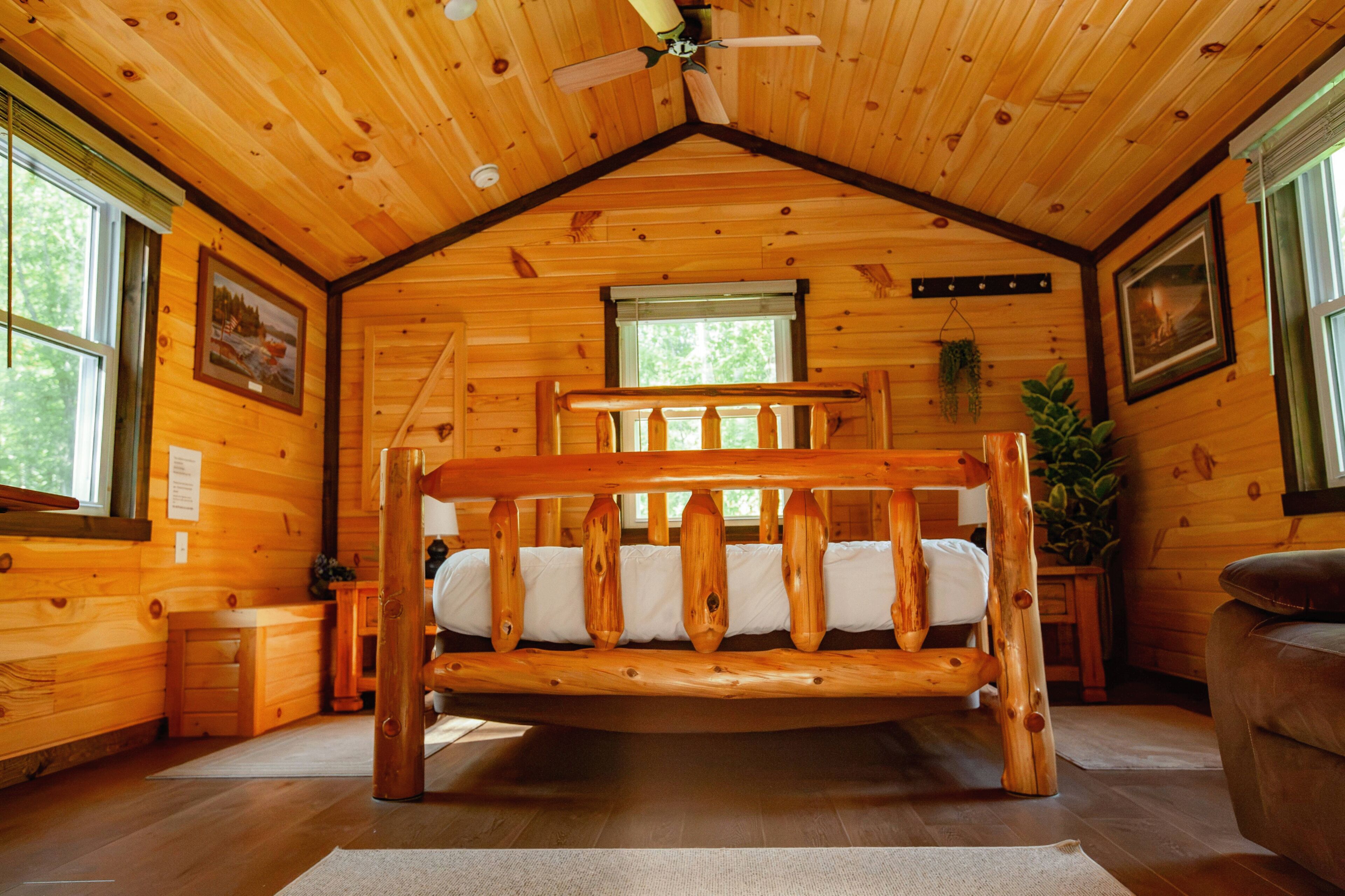 Interior d'una cabana de fusta amb un llit al centre, un ventilador de sostre i quadres a les parets.