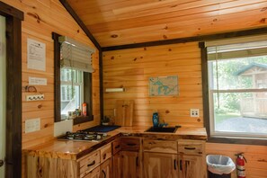 Eine Holzhütte mit einer Küche, ausgestattet mit einem Gasherd, einer Spüle, Schränken und einem Fenster mit Blick auf einen Wald.