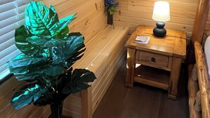A wooden cabin interior with a framed picture on the wall, a bedside table with a lamp, and a large plant.