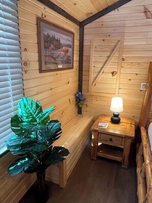 Interior de una cabaña de madera con un cuadro en la pared, una mesita de noche con una lámpara y una planta grande.