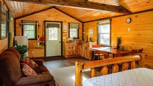 A cozy cabin interior with a living room, kitchen, and dining area, featuring wooden walls and ceilings.