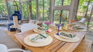 A dining area with a wooden table set for four, surrounded by large windows offering a view of greenery. - Luxurious 4BD near Downtown and RDU, with sunroom, theatre, games, & more! (Raleigh)