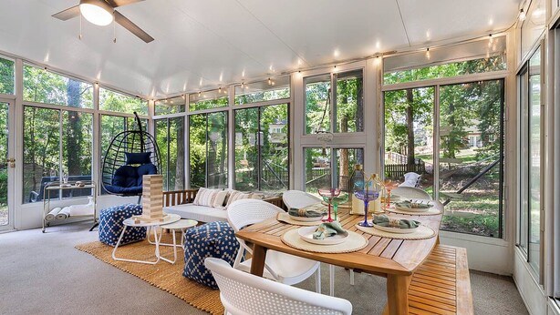 A sunroom with a wooden table, white chairs, a hanging chair, and large windows offering a view of the outdoors. - Luxurious 4BD near Downtown and RDU, with sunroom, theatre, games, & more! (Raleigh)
