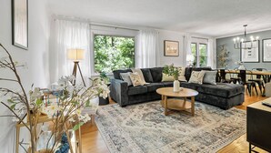 A modern living room with a dark grey sofa, a round coffee table, and a patterned rug. There is a dining area with a table and chairs visible in the background. - Luxurious 4BD near Downtown and RDU, with sunroom, theatre, games, & more! (Raleigh)
