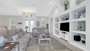 A living room with a sofa set, a coffee table, a TV on a white cabinet, and decorative items on shelves. - Cape Ohana 1505 Gulf Access Waterfront Home w/ heated Pool/ Spa & Kayaks (Cape Coral)