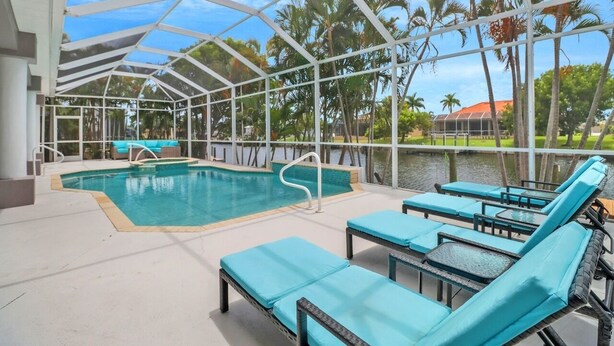 An indoor pool area with lounge chairs and a view of palm trees and water. - Cape Ohana 1505 Gulf Access Waterfront Home w/ heated Pool/ Spa & Kayaks (Cape Coral)