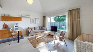 Ein modernes Wohnzimmer mit Sofa, Couchtisch und Fernseher. Es gibt eine Küchenzeile mit Schränken und einer Mikrowelle. Ein Fenster bietet einen Blick ins Freie.
