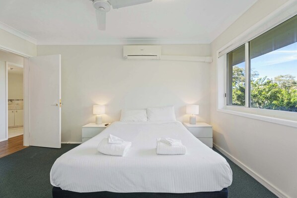 A hotel room with a large bed, two bedside lamps, an air conditioning unit, and a window with a view of trees.