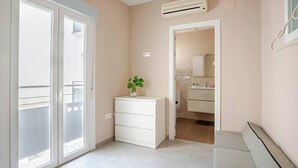 Une pièce dotée d’une porte-fenêtre coulissante, d’une commode blanche, d’une plante en pot, d’une petite salle de bain avec des toilettes et un lavabo, et d’un appareil de climatisation fixé au mur.