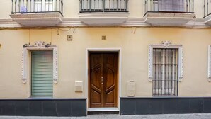 Une façade d’immeuble avec une porte en bois au centre, deux fenêtres dotées de cadres décoratifs et un balcon avec une balustrade.