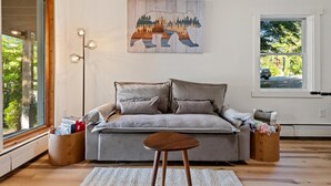 Un salón moderno con un sofá gris, un taburete de madera y un cuadro con una escena de bosque.