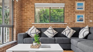 Uma sala de estar com sofá modular cinza, mesa de centro branca e janela com vista para árvores.