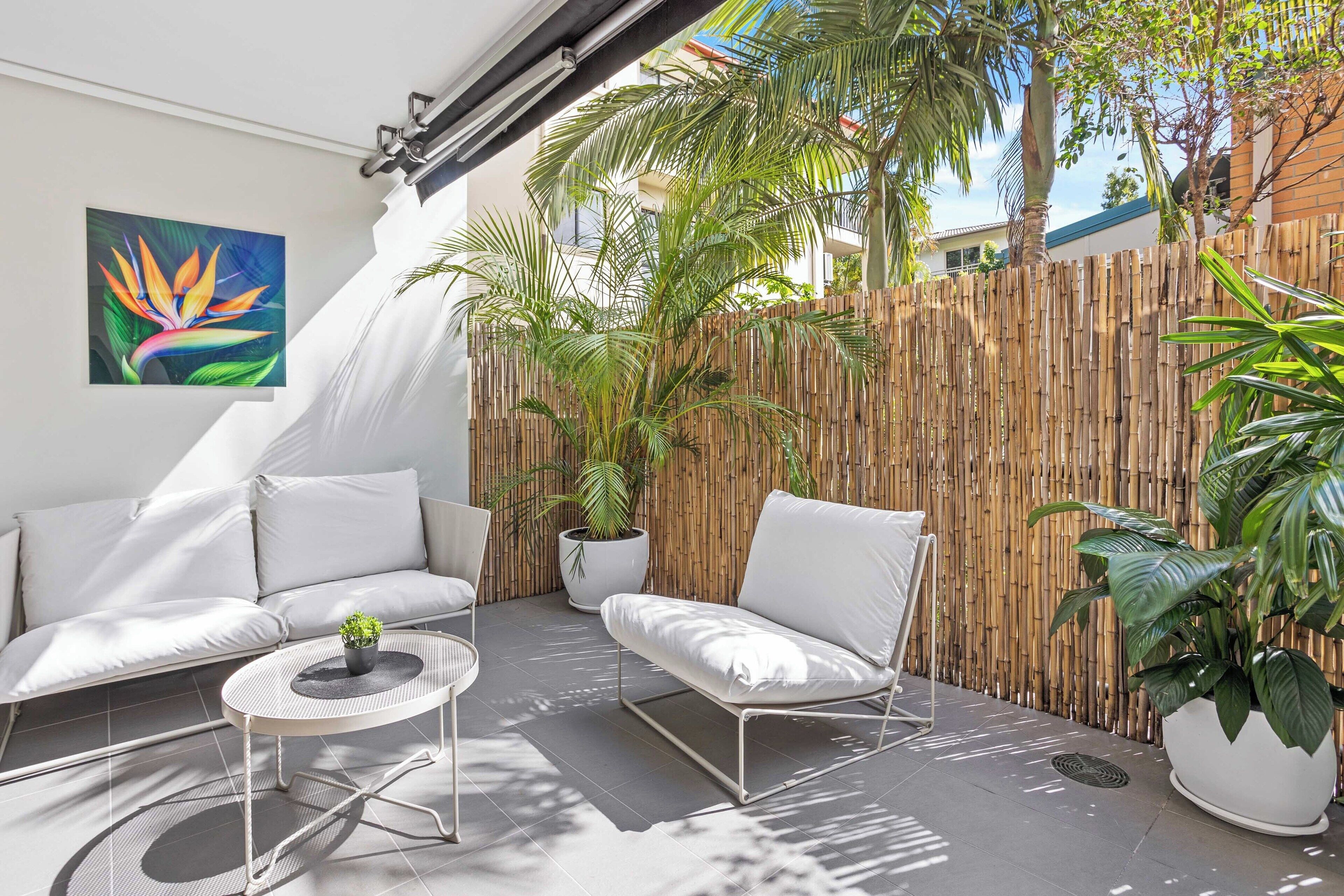 A modern outdoor patio with white furniture, a bamboo fence, and lush greenery.
