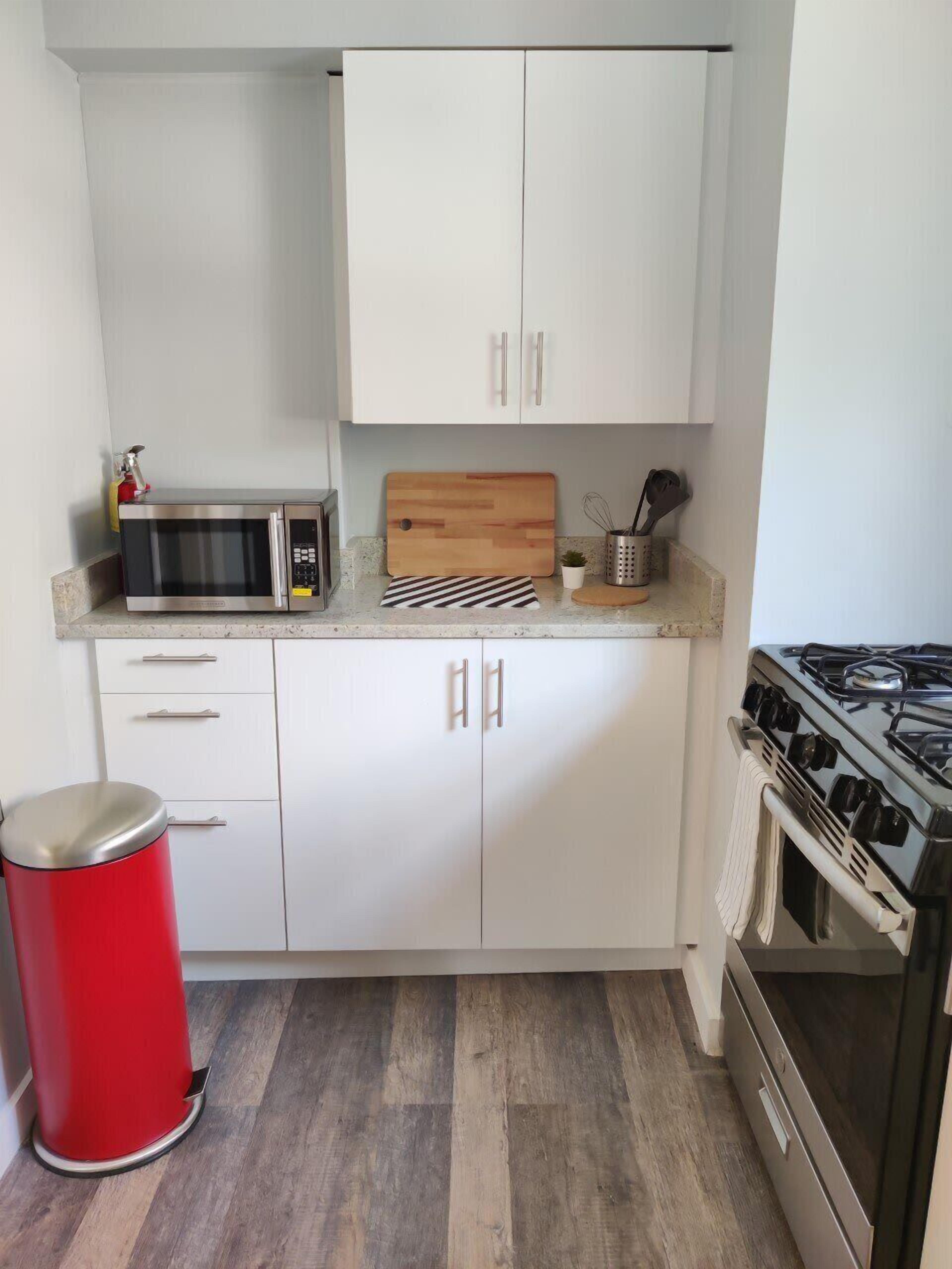 Una cocina compacta con armarios blancos, un microondas, una tabla de cortar de madera, una estufa a gas y un cubo de basura rojo.