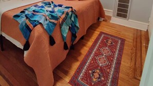 A hotel room with a bed, a window with blinds, an air conditioning unit, a nightstand, a lamp, two framed pictures on the wall, and a patterned rug on the floor. - 1601 Lombard  (Philadelphia)