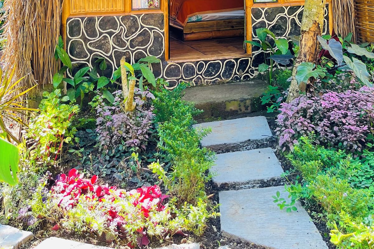 A thatched-roof hut with a bed inside, surrounded by a garden with various plants and a stone pathway leading to the entrance.