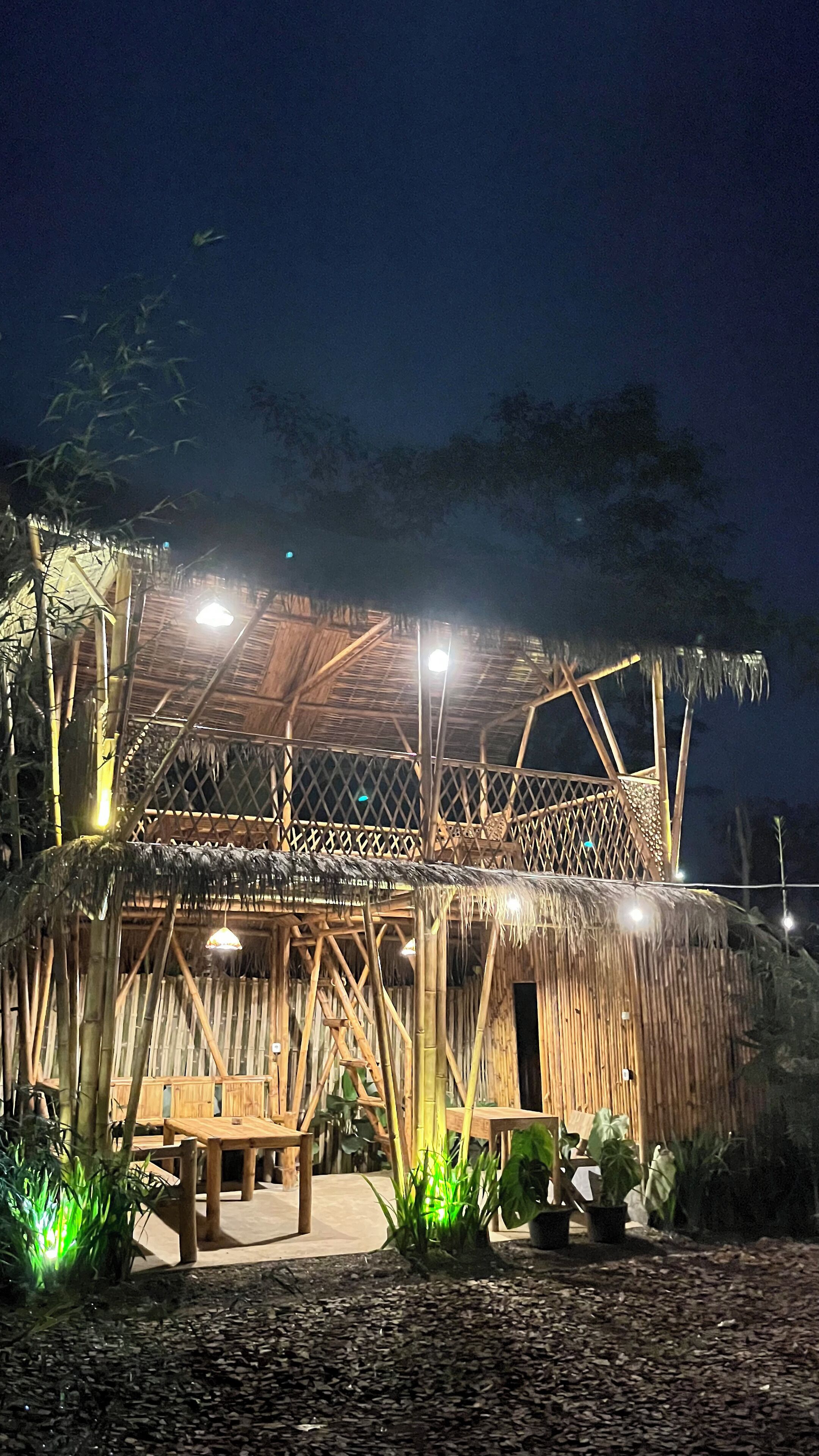 A thatched-roof structure with a wooden deck and seating area, surrounded by plants and a gravel ground.