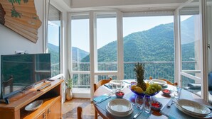 Ein Essbereich mit einem Tisch für vier Personen, einem Fernseher auf einer Holzkommode und Blick auf eine gebirgige Landschaft durch große Fenster.