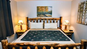 A bed with a wooden frame, two bedside tables with lamps, a painting on the wall, and a quilt on the bed. - One Bedroom, Two Bath Condo with River View (Estes Park)