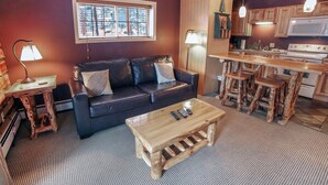 A cozy living room with a leather sofa, wooden coffee table, and a kitchen area with wooden stools and a microwave.