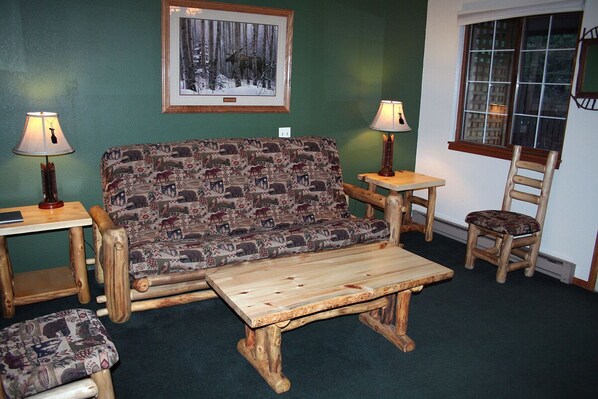 Un cuarto acogedor con un sofá estampado, una mesa de centro de madera, dos lámparas, una silla y un cuadro en la pared.