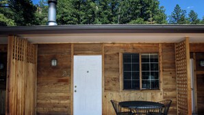 A wooden cabin with a white door, a window with a grid pattern, and a patio with a table and chairs. - Studio with Full Kitchen, Queen bed, Patio with Personal Grill (Estes Park)