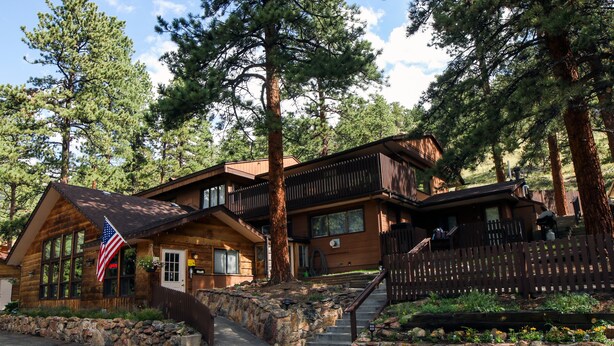 A log cabin with a stone staircase, wooden deck, and a flag. - Studio with Full Kitchen, Queen bed, Patio with Personal Grill (Estes Park)