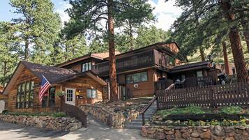 A log cabin with a stone staircase leading to a wooden deck, surrounded by tall pine trees.