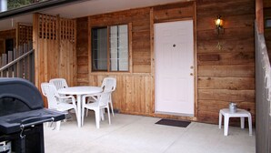 Una cabaña de madera con puerta blanca, un letrero con el número 4, una parrilla y una mesa con sillas.