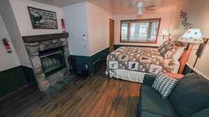 Ein Zimmer mit Kamin aus Stein, einem Bett mit Steppdecke, einer Couch, einem Fenster mit Jalousien und einem Deckenventilator.
