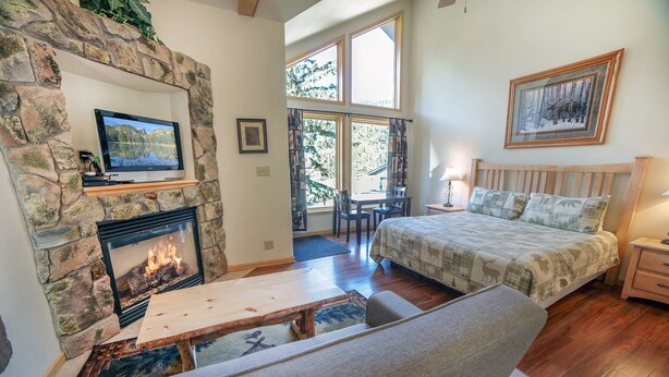 A cozy room with a stone fireplace, a bed with a forest-themed bedspread, a desk, and a window with a view of trees. - Deluxe Spa Suite, jetted tub for 2, fireplace (Estes Park)