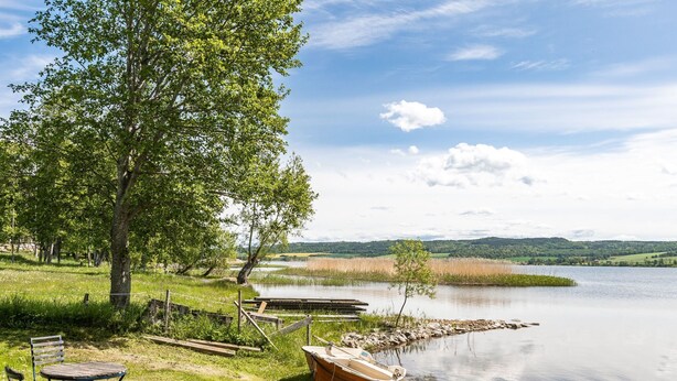 A lakeside setting with a wooden boat, a metal table and chairs, and a grassy area with trees.