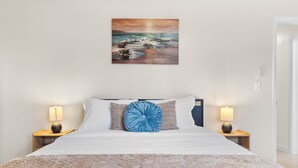 Een net opgemaakt bed met een beige deken, twee bedlampen en een schilderij van een strand.