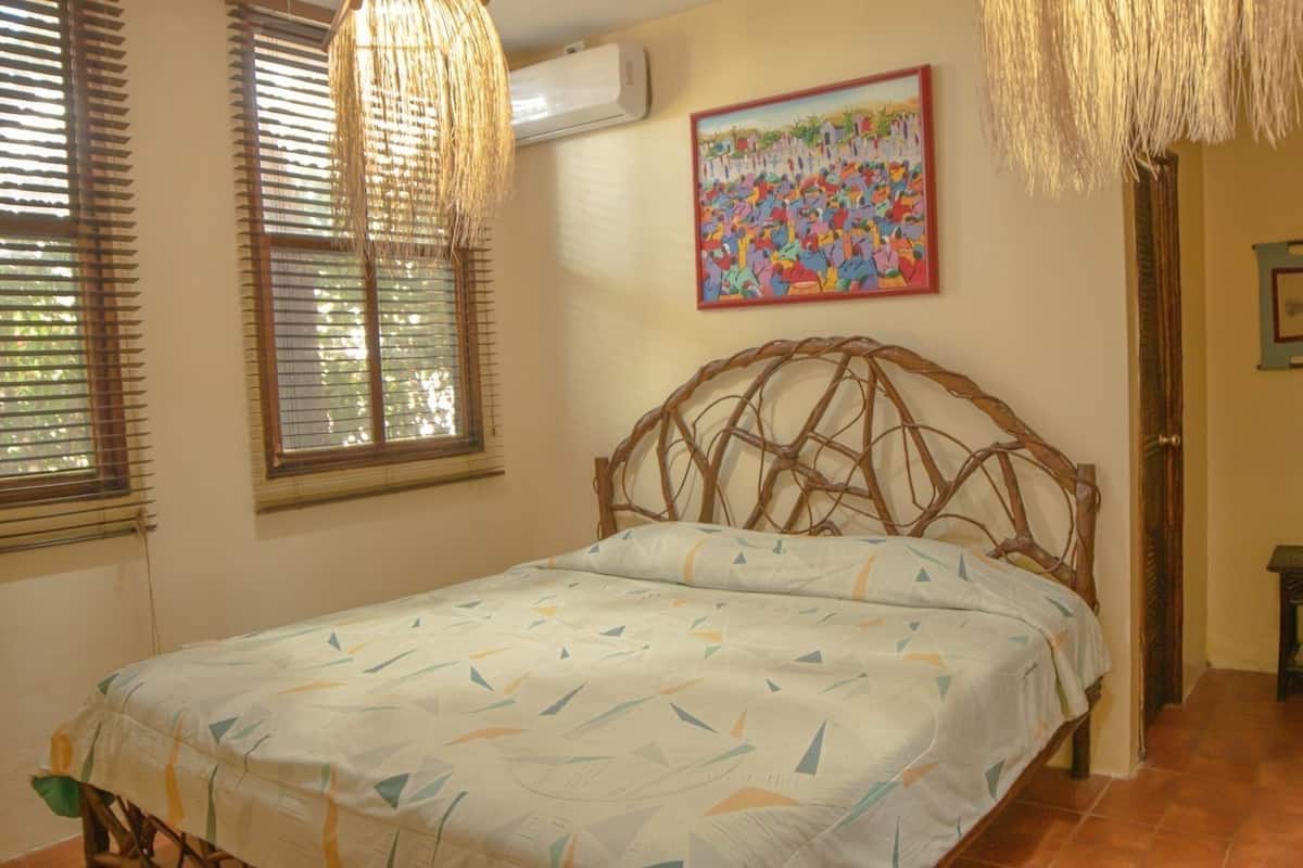 A bedroom with a bed, a window with blinds, a wall-mounted air conditioner, a colorful painting, and a small table.