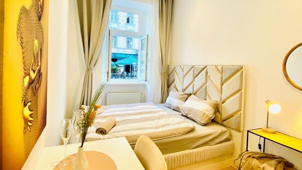 A hotel room with a bed, a bedside table with a lamp, a window with curtains, and a view of the street outside. - Ottakring Gem 4-BR Retreat/Breathtaking City Views (Vienna)