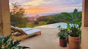 A balcony with lounge chairs, potted plants, and a view of the ocean at sunset.