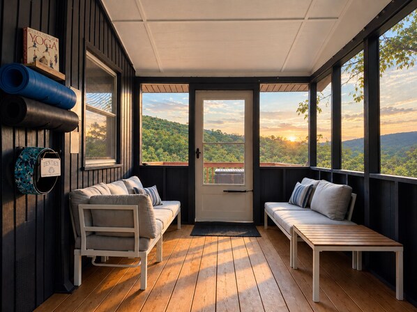 A cozy room with a wooden floor, a white sofa, a bench, and a view of a forested landscape through large windows.