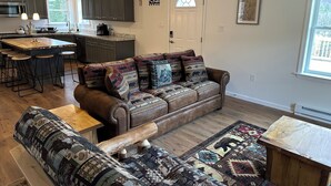 A cozy living room with a brown leather sofa and matching ottoman, a wooden coffee table, and a patterned rug. A kitchen with dark cabinets and a large island is visible in the background. - ADK cottage with Mountain Views  (Saranac)