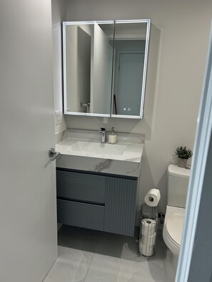 A modern bathroom with a white vanity, a large mirror with integrated lighting, a toilet, and a small potted plant. -  Breathtaking view, as well as modern, clean & well-appointed apartment 
 

 (Toronto)