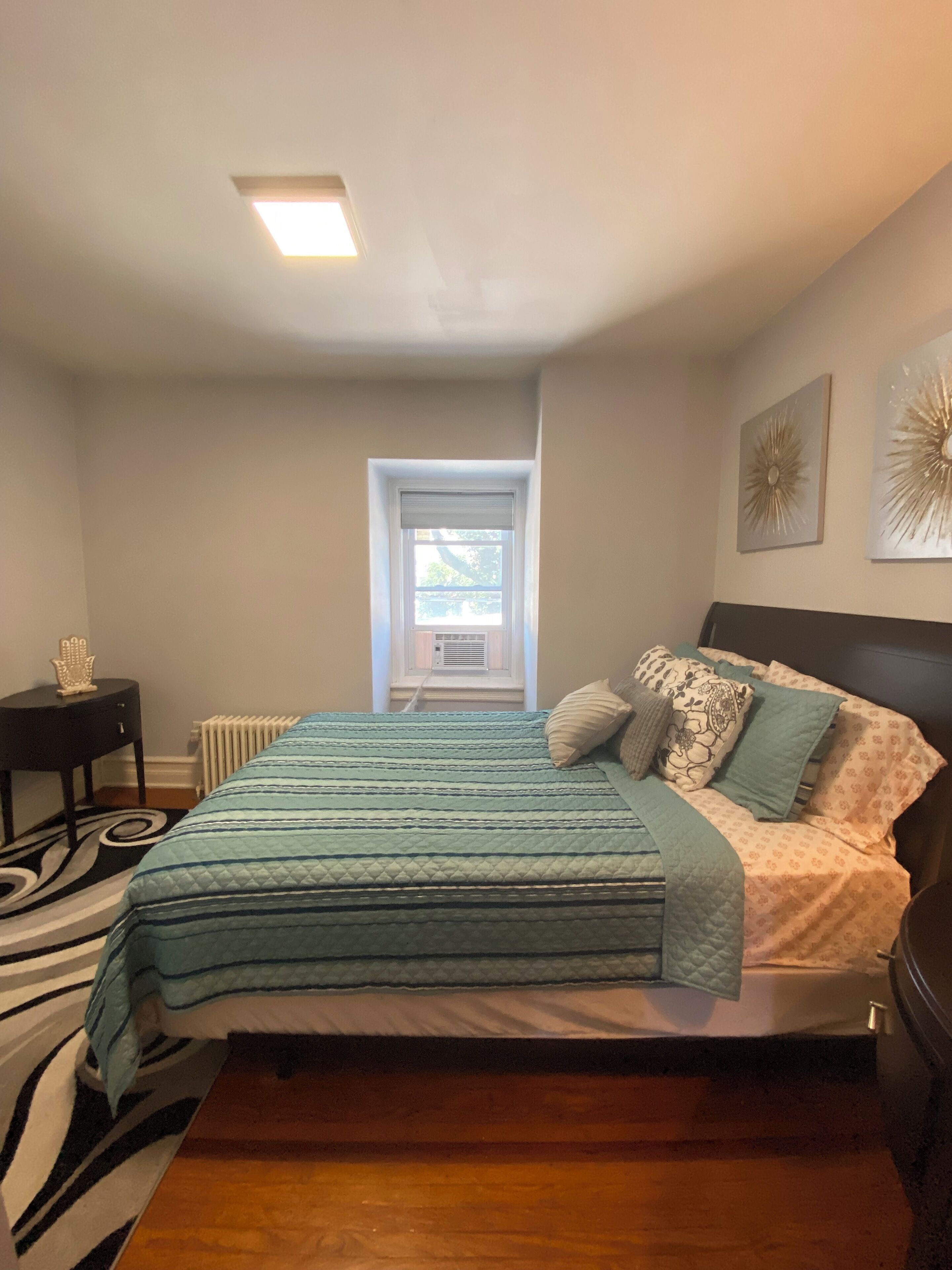 A bedroom with a bed, a nightstand, a radiator, a window, and a ceiling light.