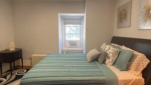 A bedroom with a bed, a nightstand, a radiator, a window, and a ceiling light.