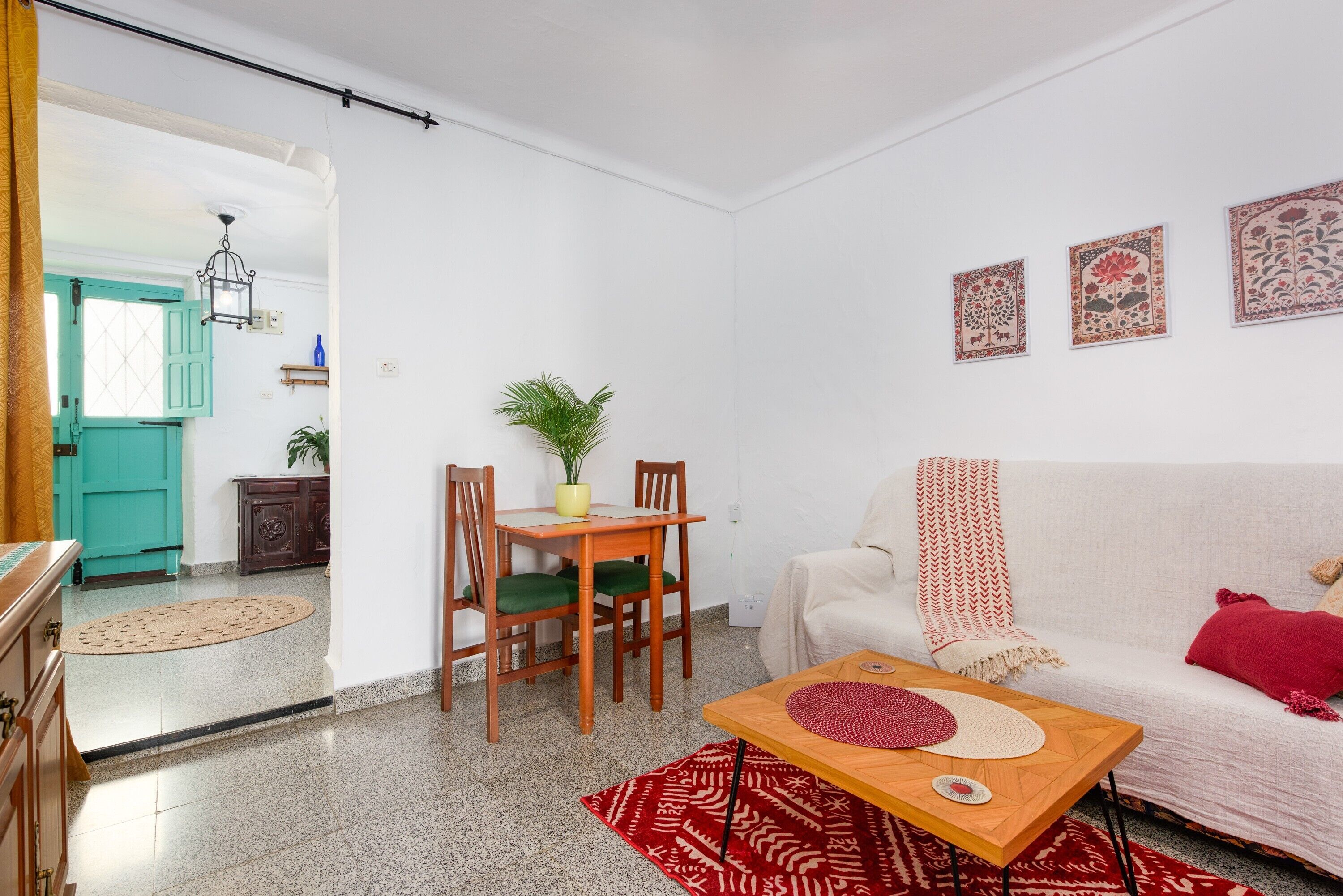 A living room with a sofa, a coffee table, a dining table with chairs, a potted plant, and a kitchen visible through an open door.
