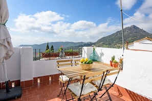 A terrace with a wooden table, chairs, a bowl of fruit, potted plants, and a view of mountains. - Holiday Home 'La Casa De Eva' with Mountain View, Shared Terrace and Wi-Fi (Gaucín)