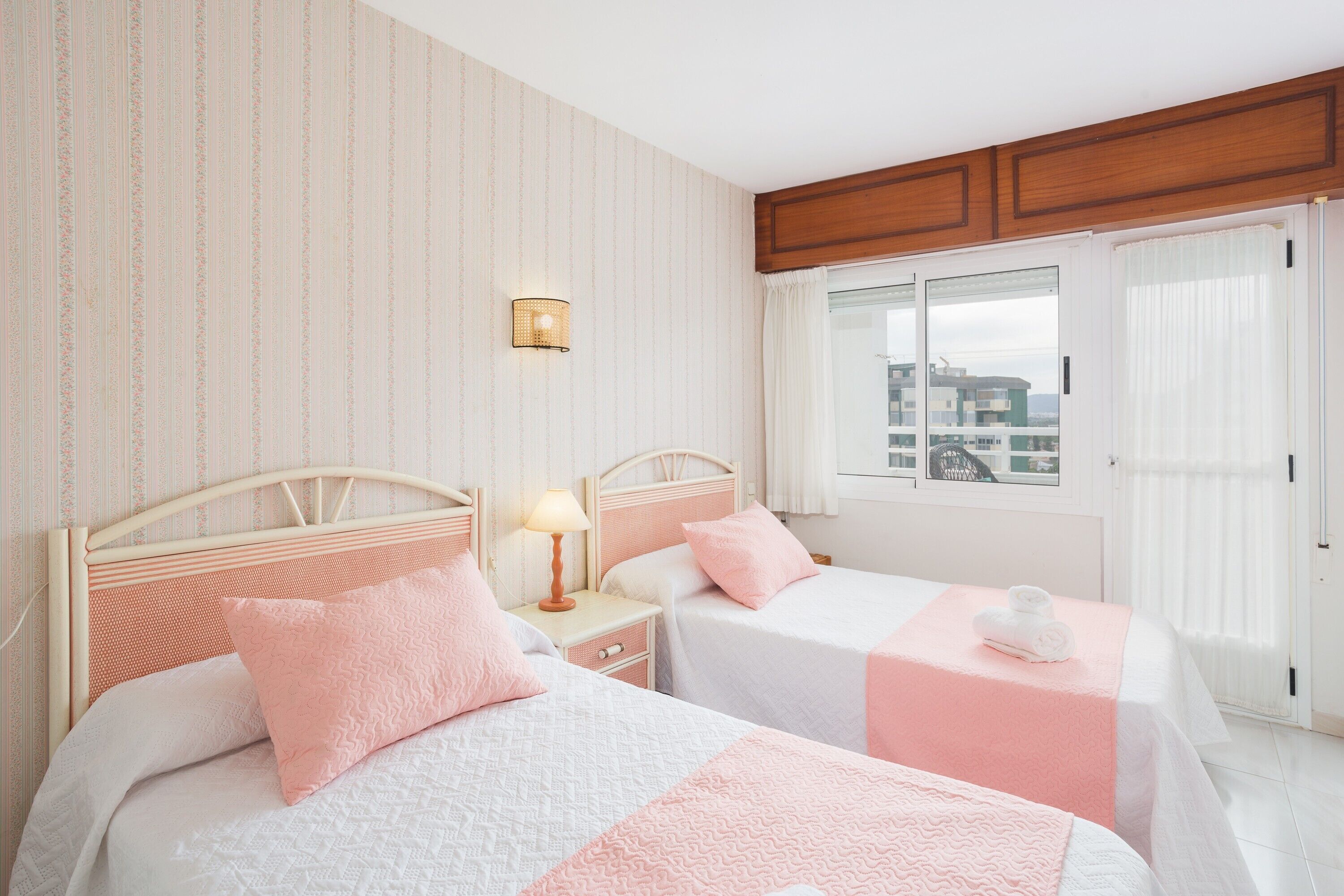 A hotel room with two beds, a nightstand with a lamp, a window with a view, and a wooden headboard.