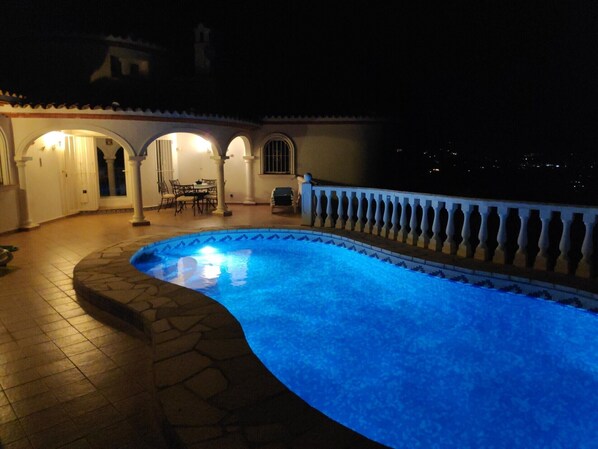 A nighttime swimming pool with a curved edge, surrounded by a tiled patio and a railing.