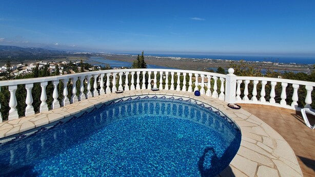 Une piscine dotée d’une balustrade blanche, entourée d’une terrasse en pierre, et offrant une vue sur un paysage côtier.